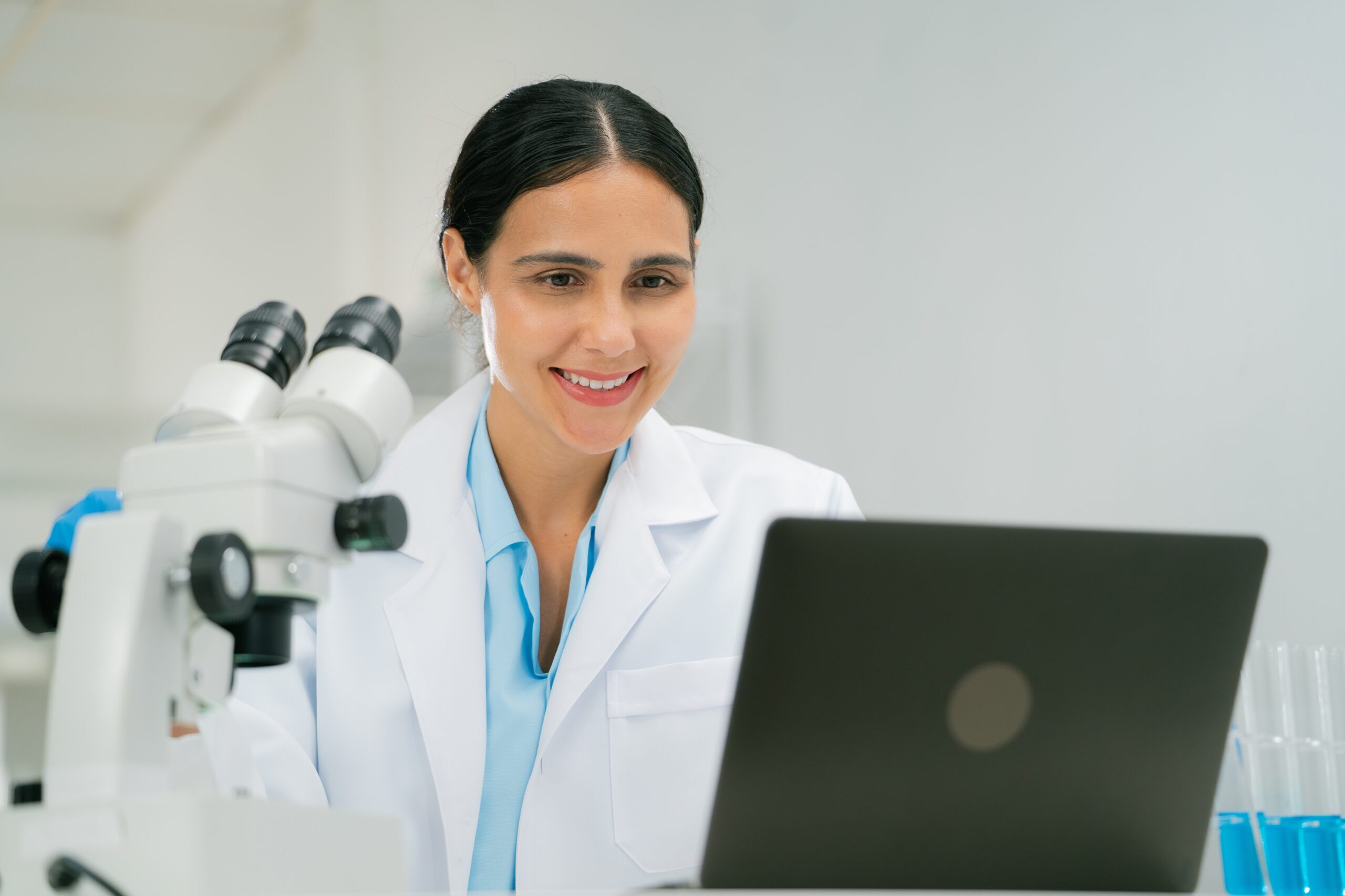scientist and researcher in laboratory setting smiles while working on laptop, with microscope and test tubes nearby, showcasing chemistry research. scientist researcher chemistry laboratory